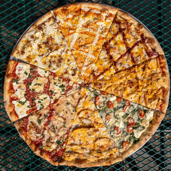 A large Tommyboy's Pizza with eight different specialty slices arranged in a circle on a green metal table.