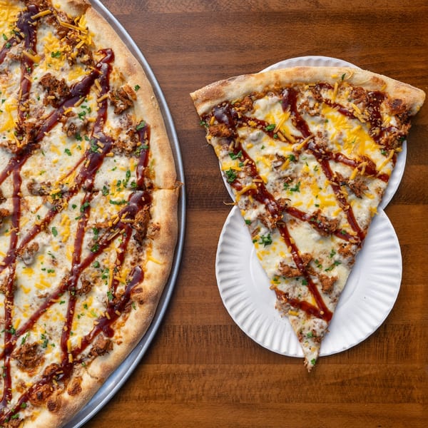 A whole BBQ chicken pizza and a single slice on a paper plate, on a wooden table.