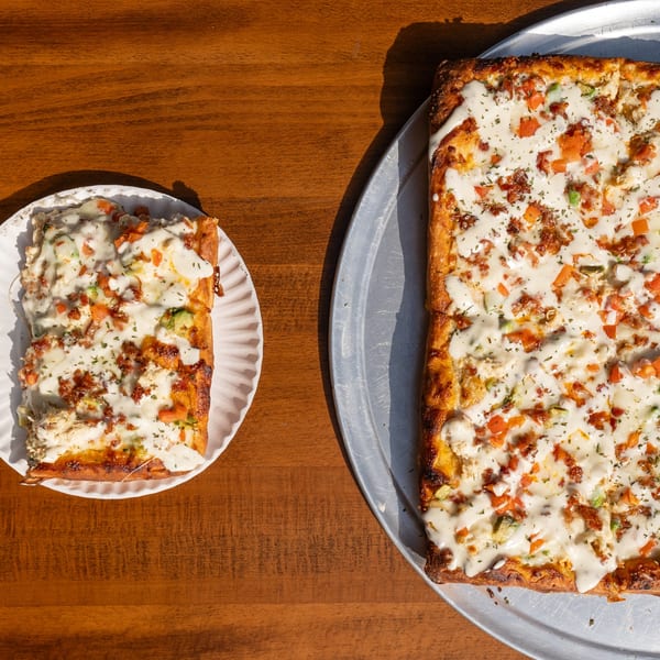 A square Tommyboy's Pizza with white sauce, bacon, and vegetables, with one slice on a paper plate.